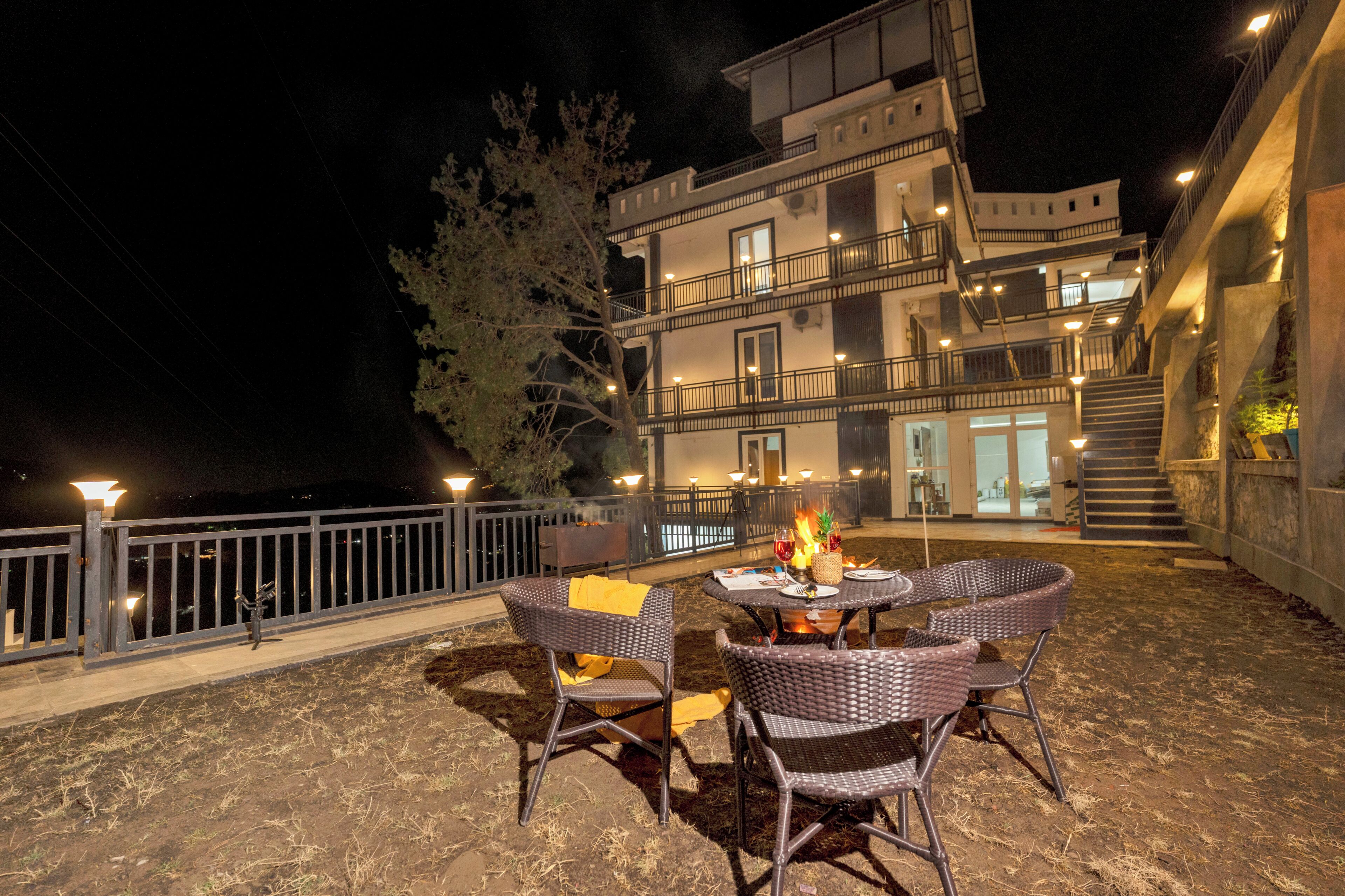 An outdoor patio with wicker chairs and a table, a fire feature, and a building with multiple balconies and stairs in the background.
