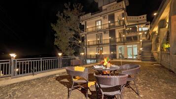 An outdoor patio with wicker chairs and a table, a fire feature, and a building with multiple balconies and stairs in the background.