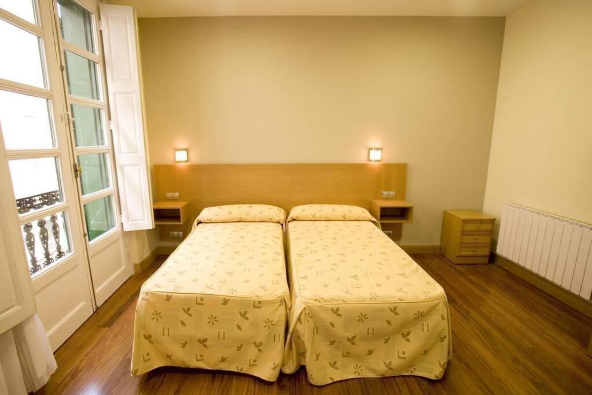 Habitación doble con dos camas, cabecero de madera, lámparas de noche, una ventana con cortinas y un radiador.