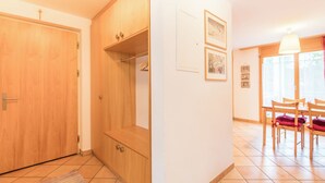 Una stanza con un'armadio in legno, un piccolo ripiano, una porta, un pavimento piastrellato, un tappeto, un'area pranzo con sedie e un tavolo, e quadri appesi al muro.