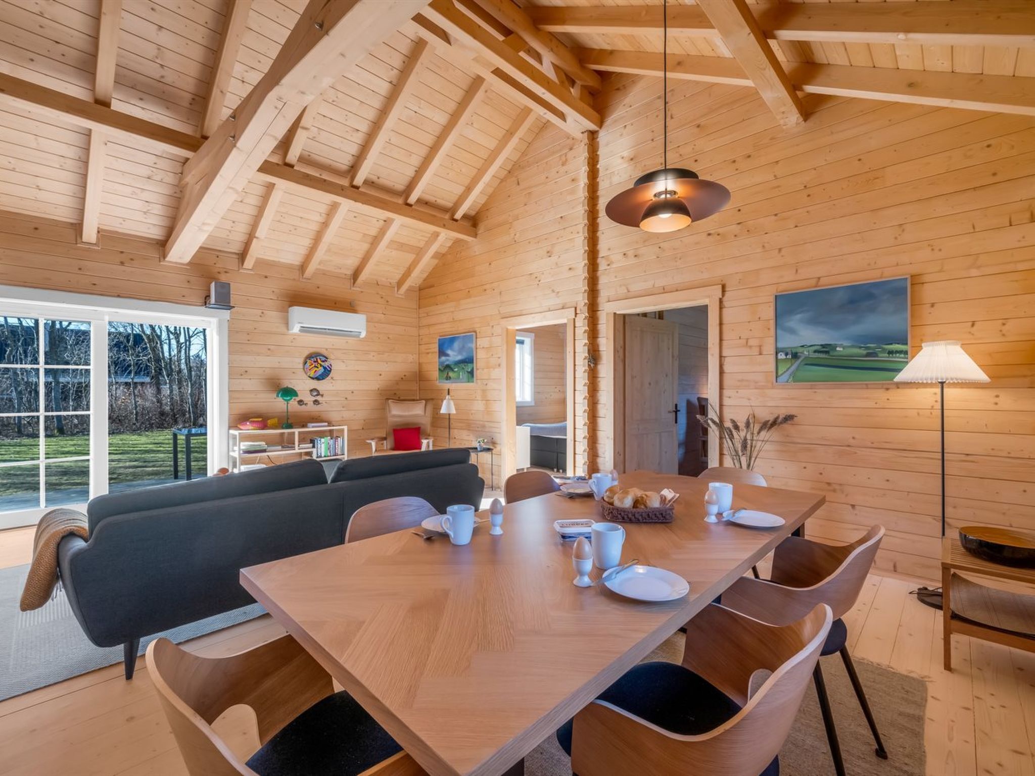 A wooden interior with a dining table set for four, a sofa, and a kitchen area with a view.