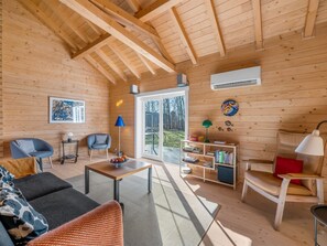 Interior de madeira com sofá, cadeiras, mesa, estante de livros e vista por uma porta de vidro.