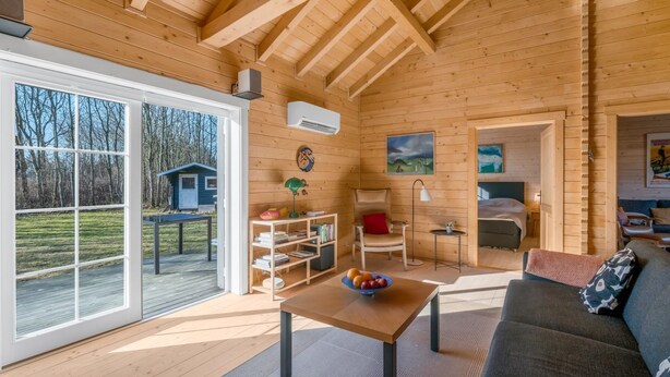 Een houten hut van binnen, met een eethoek, een bank, een tafel met een schaal fruit, en een schilderij aan de muur.