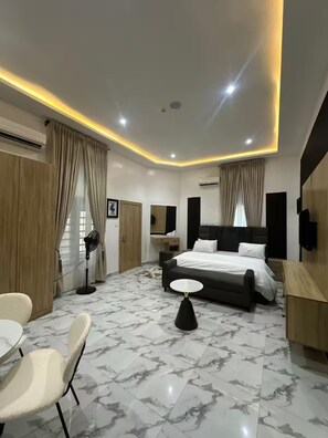 A modern hotel room with a bed, a desk, a chair, a fan, and a TV. - 4bedroom Luxury Apartment (Lagos)