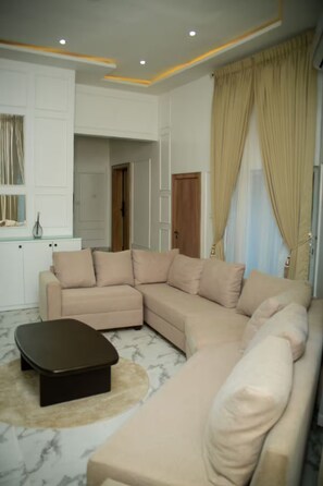 A living room with a beige sectional sofa, a black coffee table, and a marble floor. - 4bedroom Luxury Apartment (Lagos)