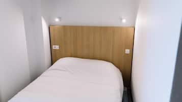 A small, narrow room with a wooden headboard, a white bed, and two wall-mounted light switches.