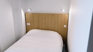 A small, narrow room with a wooden headboard, a white bed, and two wall-mounted light switches. - Welcomotel (Limoges)