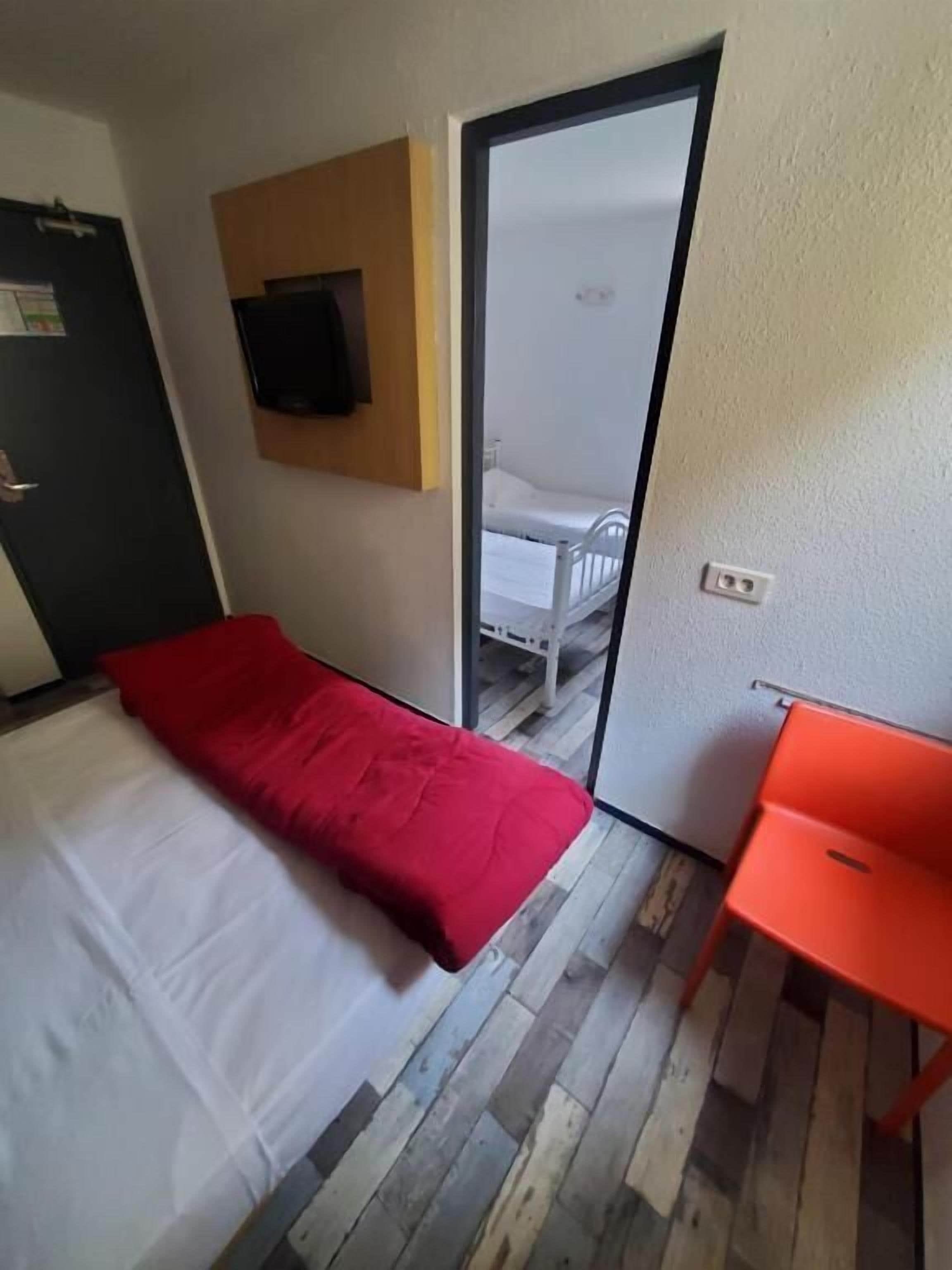 A room with a bed, a red chair, a wooden floor, a television on the wall, and a door leading to another room.