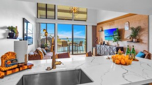 A modern living room with a large window overlooking the sea, a kitchen with a marble countertop, and a dining area with a view. - Napili Point 2 Oceanfront Villas, Sleeps 8 | Car Incl. w/ 6 nights | ML-4106 by KBM (Lahaina)