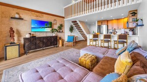 A modern living room with a large TV, wooden flooring, a staircase, and a kitchen area. - Napili Point 2 Oceanfront Villas, Sleeps 8 | Car Incl. w/ 6 nights | ML-4106 by KBM (Lahaina)