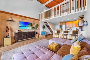 Ein modernes Wohnzimmer mit großem Fernseher, Holzboden, einer Treppe und einer Küchenzeile.