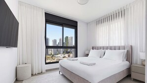 Un moderno cuarto de hotel con una cama grande, vistas a la ciudad, un otoman redondo y una mesita de noche con lámpara.