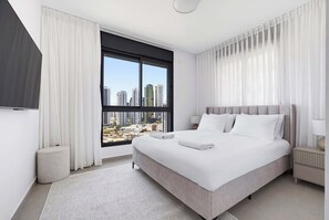 Ein modernes Hotelzimmer mit einem großen Bett, Blick auf die Stadtlandschaft, einem runden Hocker und einem Nachttisch mit Lampe.