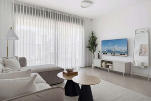 Ein modernes Hotelzimmer mit Sofa, rundem Couchtisch, Fernseher und großem Fenster mit leichtem Vorhang.