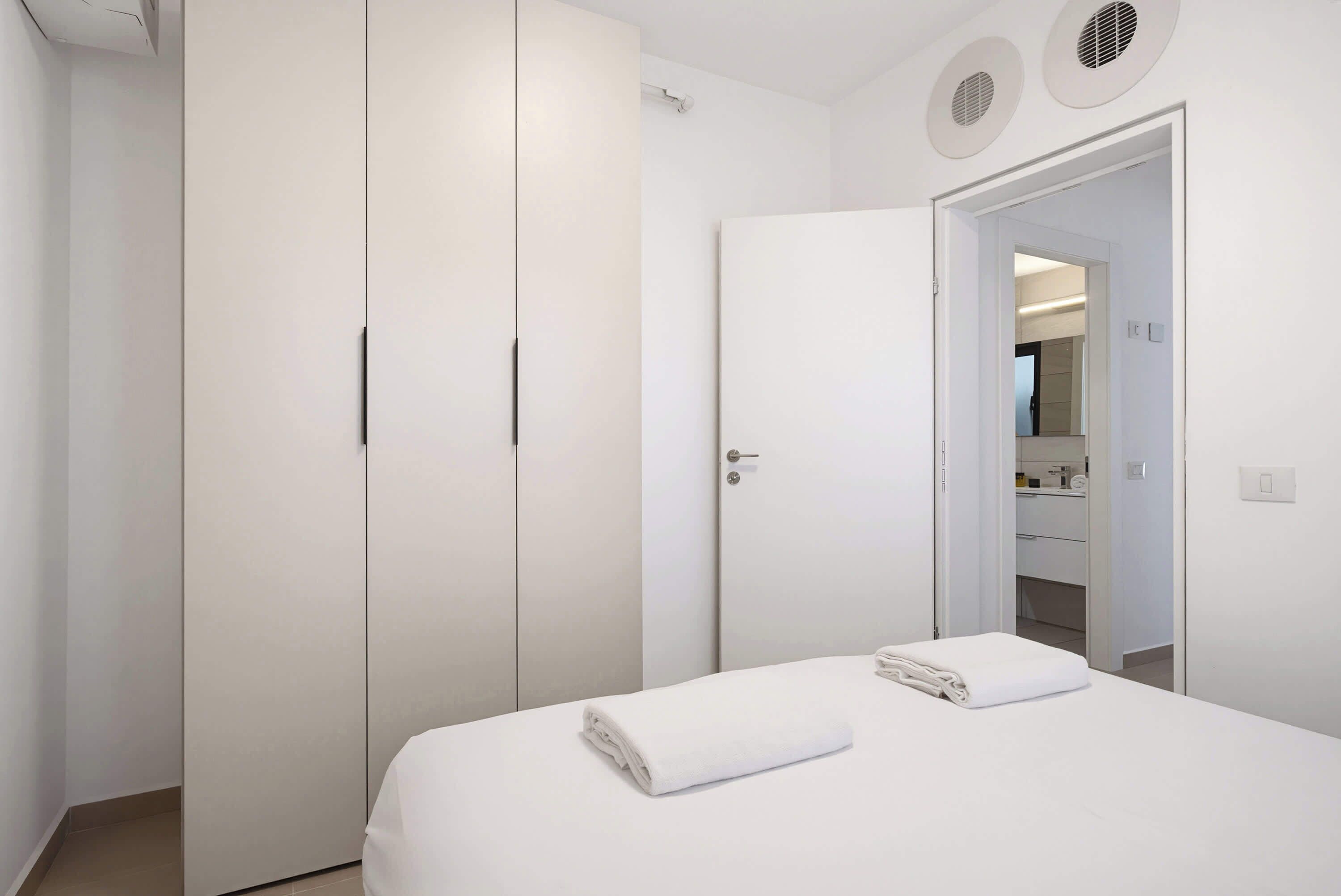 A modern hotel room with a large wardrobe, a bed with neatly folded white towels, and a bathroom visible through an open door.