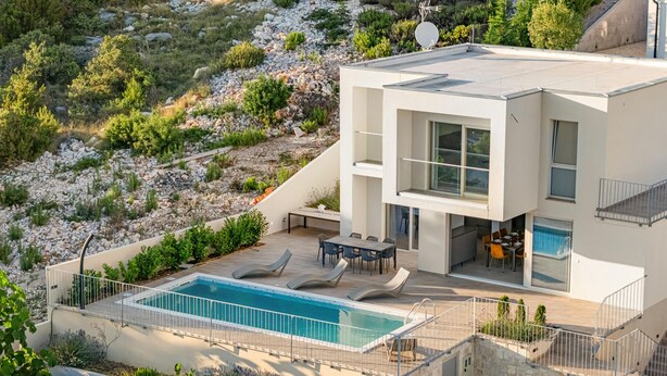 Ein modernes, zweistöckiges Haus mit Pool, Sitzgelegenheit im Freien und Blick auf einen felsigen Hügel.