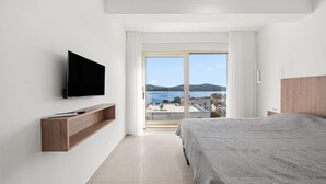 Ein Hotelzimmer mit einem Bett, einem an der Wand befestigten Fernseher, einer Holzablage und einem großen Fenster mit Blick auf das Meer und Gebäude.