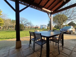 Eine überdachte Terrasse mit Tisch und Stühlen, ein geparktes Auto und eine Rasenfläche mit Bäumen und Gebäuden im Hintergrund.