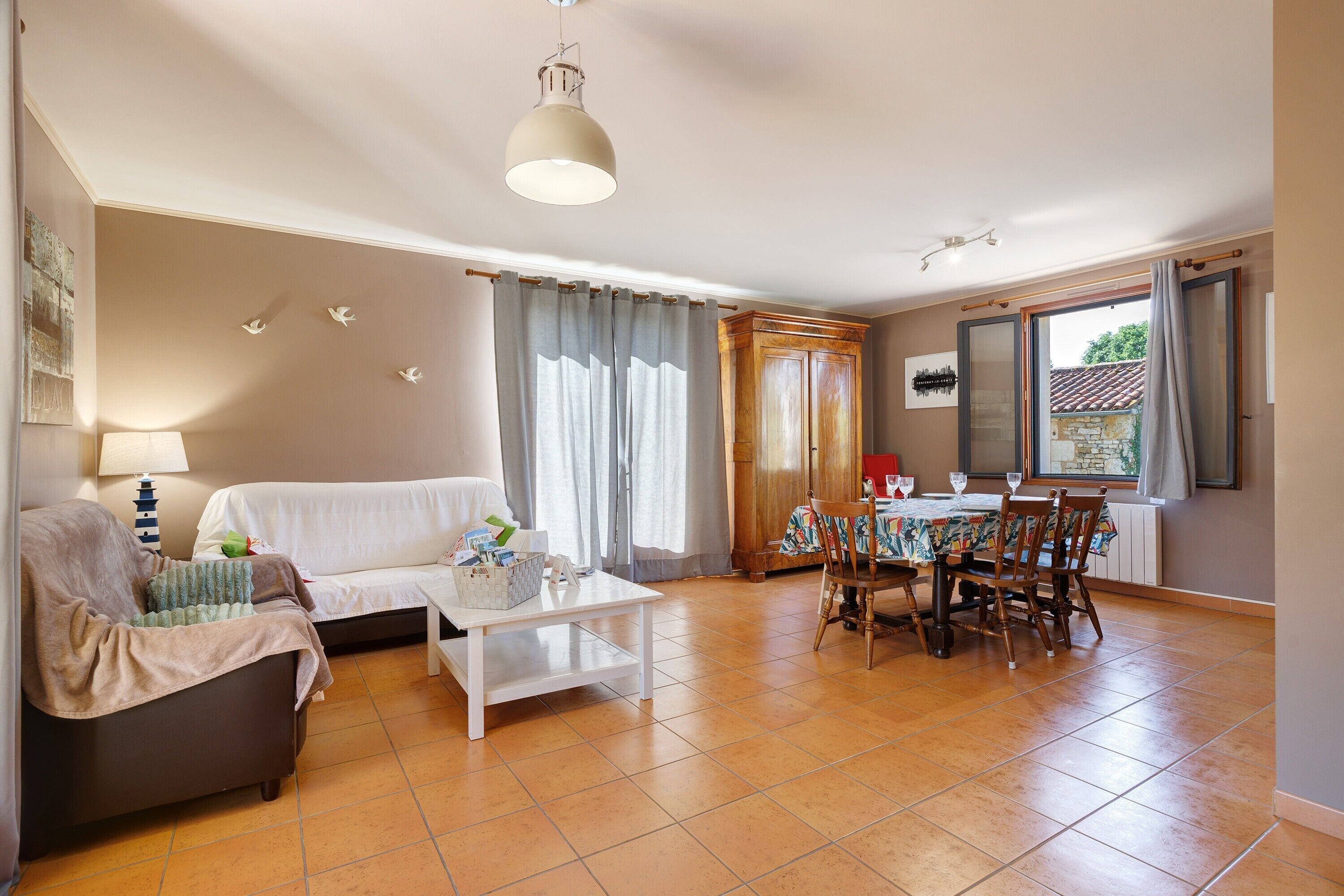 Un salon comprenant un canapé, une table basse, une table à manger avec des chaises, une lampe et une fenêtre avec des rideaux.