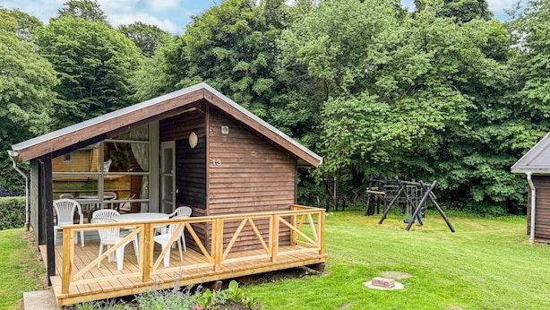A wooden cabin with a deck, a table with chairs, and a swing set in a grassy area.