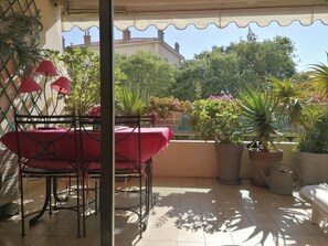 Eine überdachte Terrasse mit Tisch und Stühlen, Blumentöpfen und Blick auf ein Gebäude im Hintergrund.