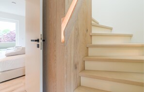 Un interior moderno con una escalera de madera, una cama con ropa blanca y un ventanal con vistas a árboles.