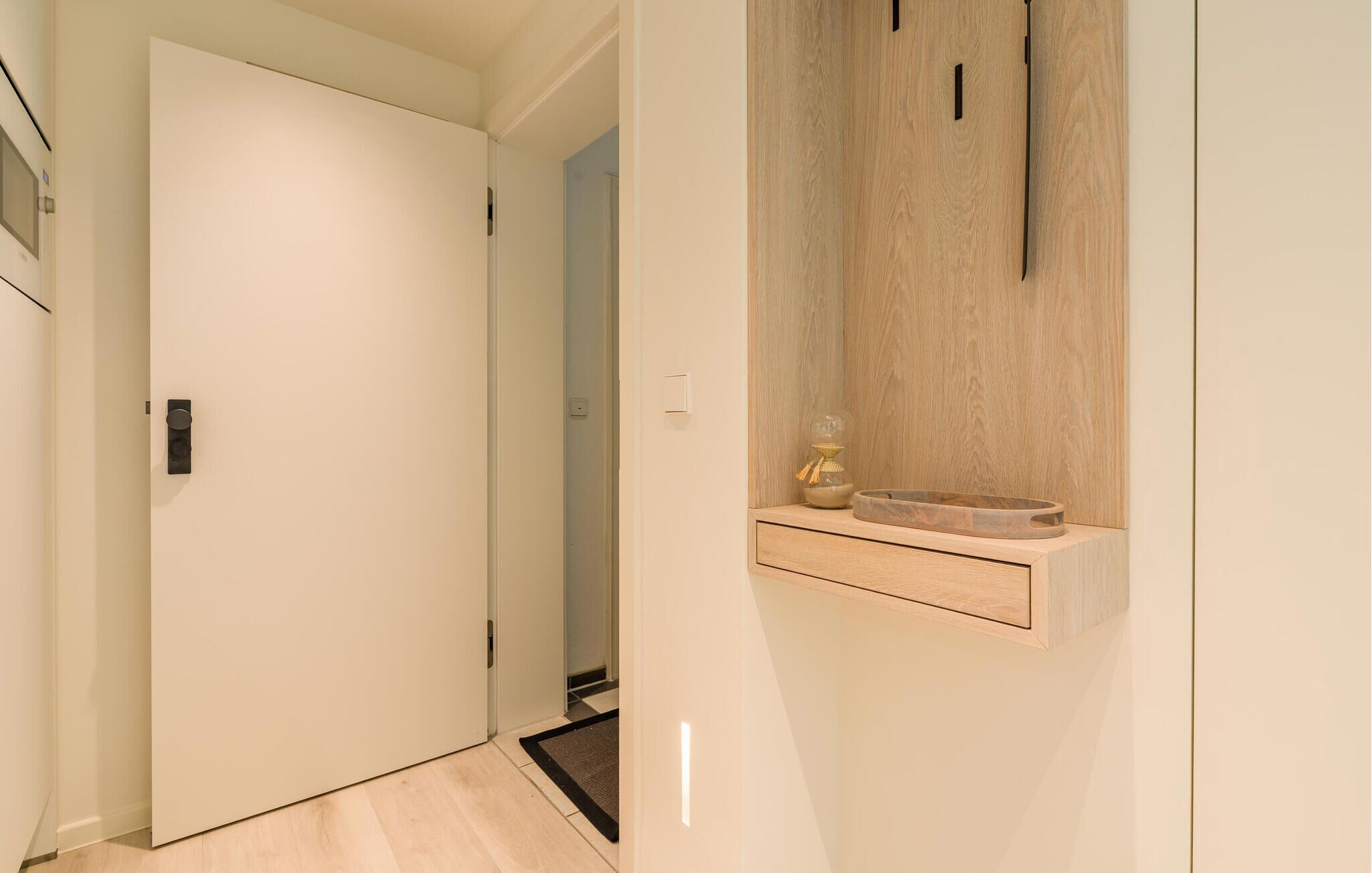 Un couloir avec une porte blanche, une étagère en bois sur laquelle se trouvent un bol et un objet décoratif, et un interrupteur.