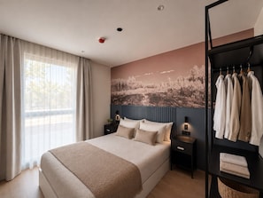 A modern bedroom with a large bed, a headboard with vertical lines, a wall-mounted shelf with clothes, and a window with curtains.