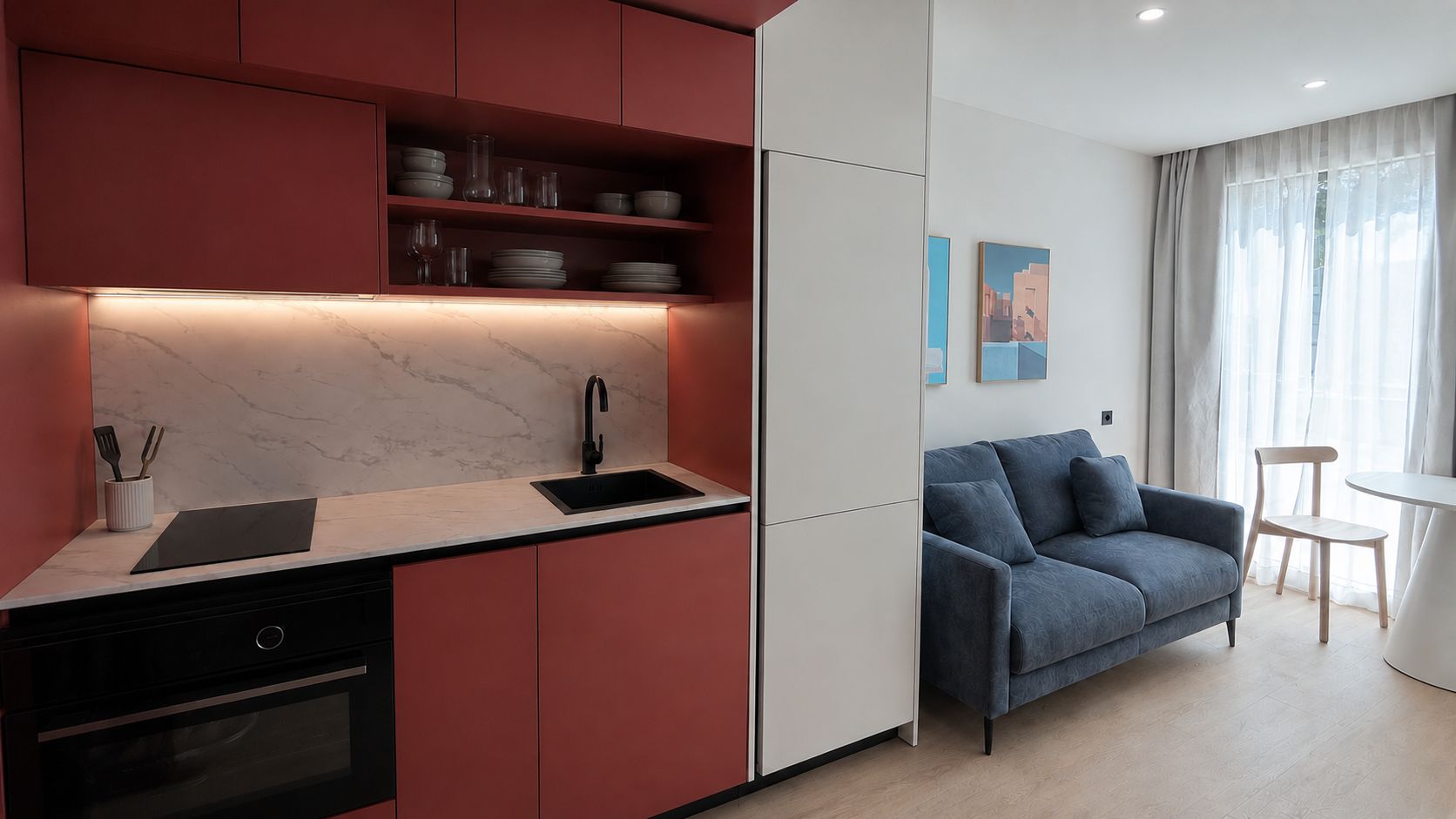 Una cocina moderna con horno empotrado, un lavabo y una encimera. En la zona de estar contigua hay un sofá azul y una mesita.
