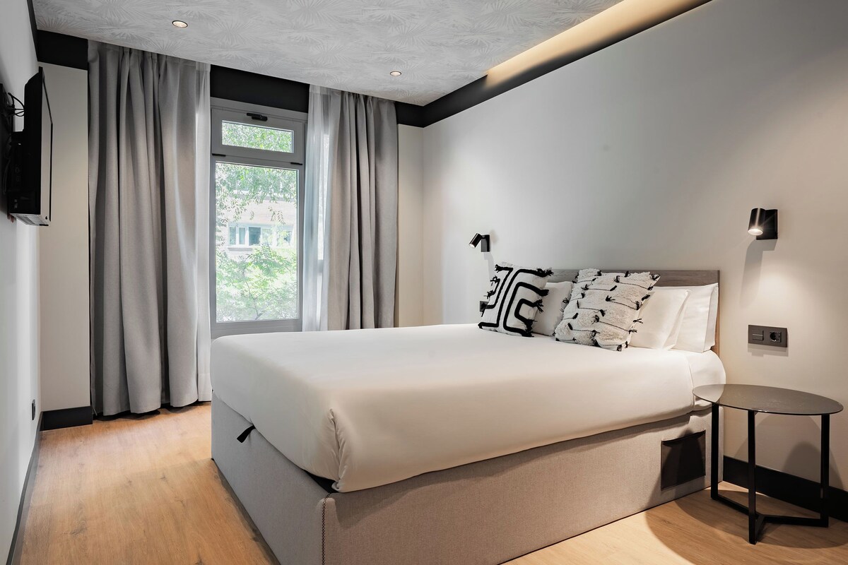 A modern bedroom with a large bed, a bedside table, and a window with curtains.