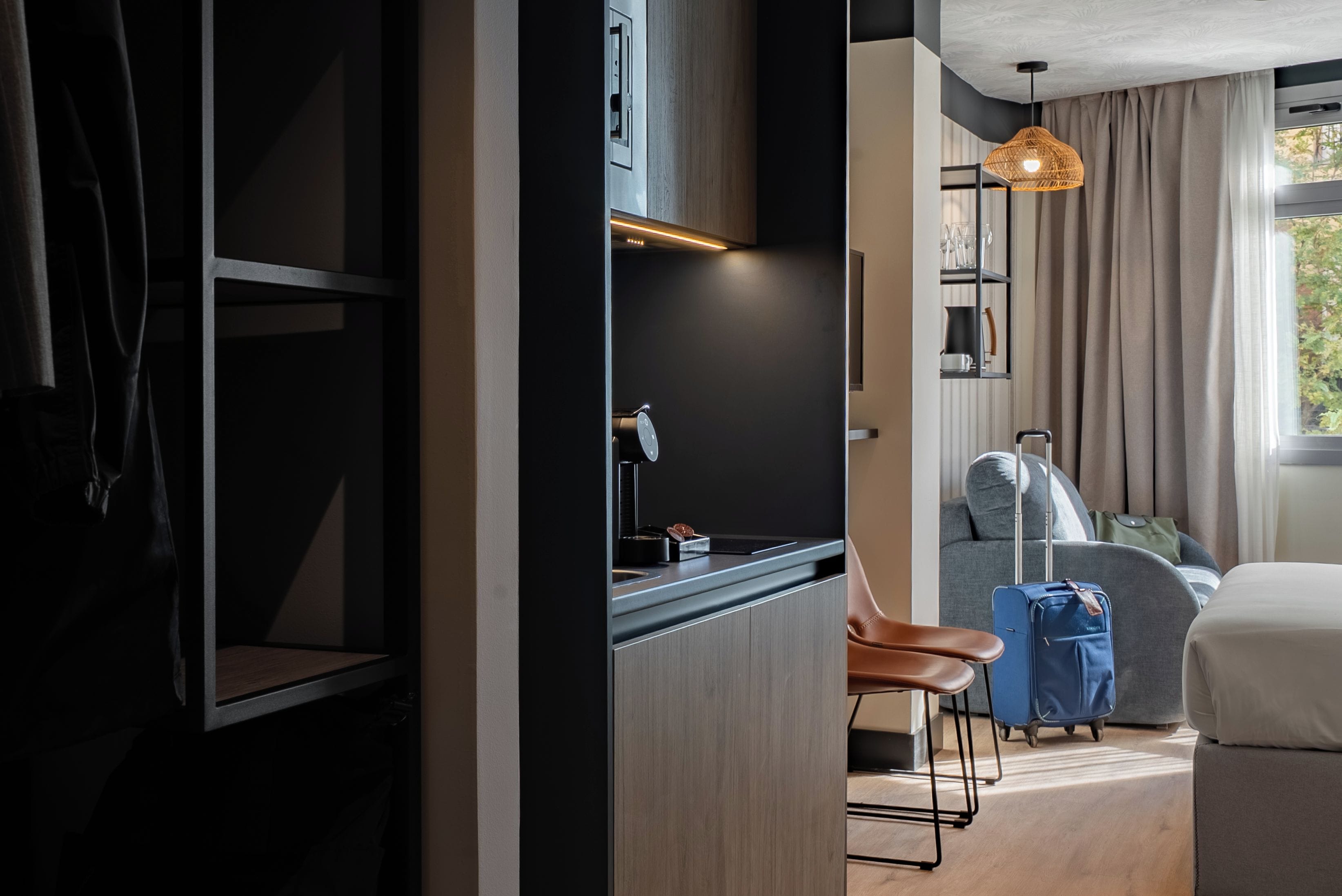 A modern hotel room with a kitchenette, a seating area with a sofa and chairs, a suitcase, and a window with curtains.