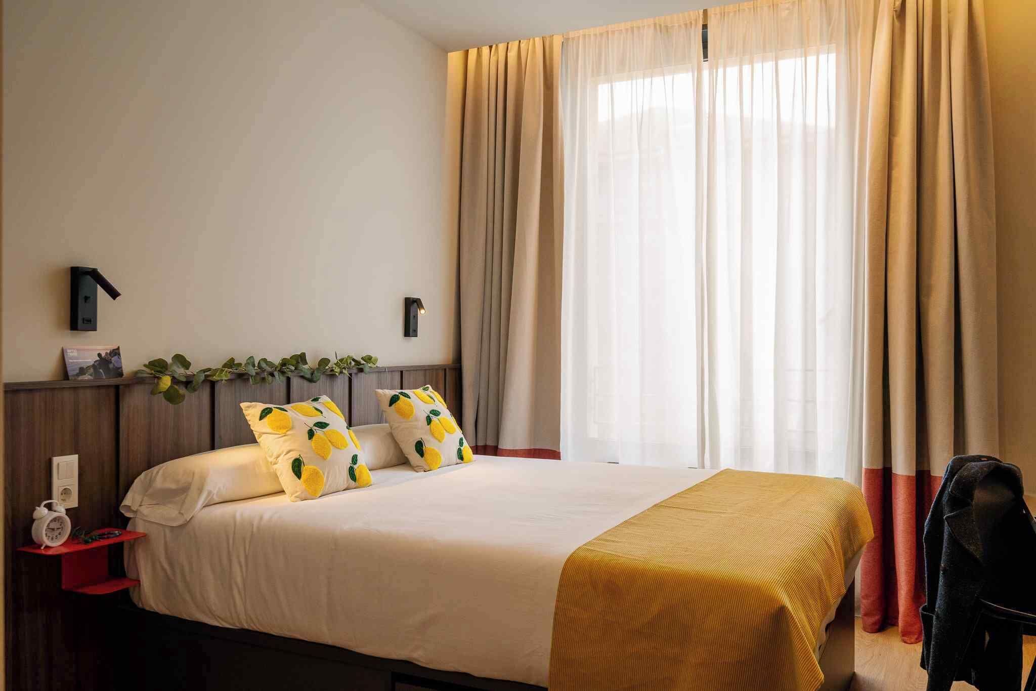 Una habitación de hotel con cama, una mesita de noche con lámpara, una silla y una ventana con cortinas.