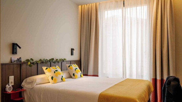 Una habitación de hotel con cama, una mesita de noche con lámpara, una silla y una ventana con cortinas.