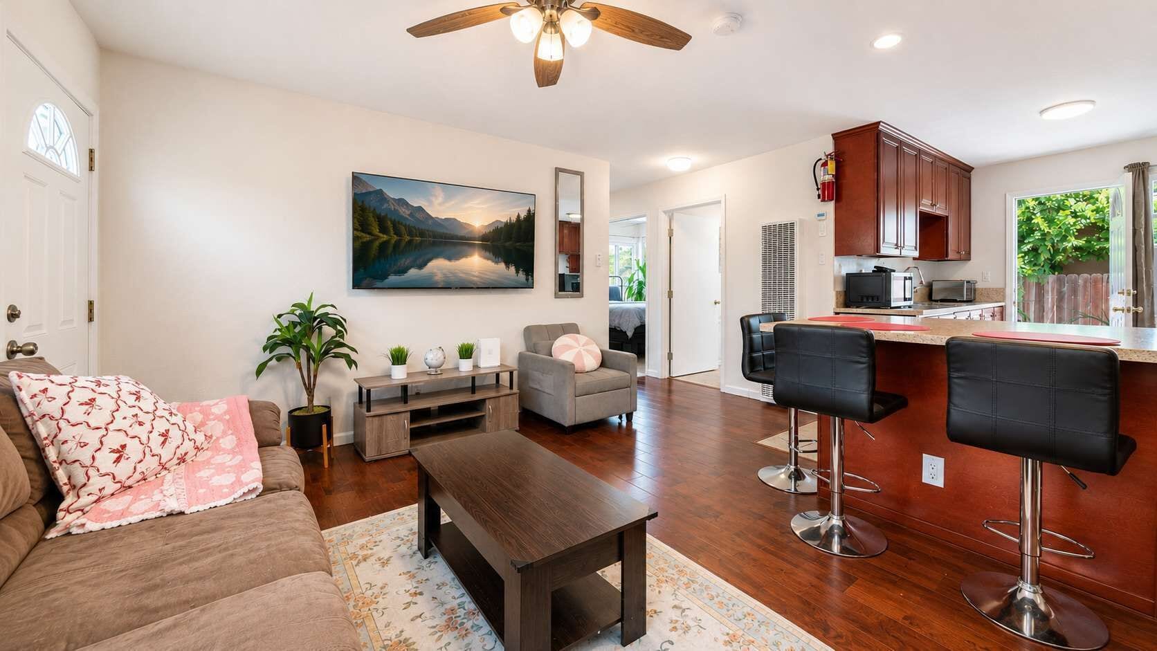 A living room with a sofa, coffee table, TV, and a painting. There is a kitchen with a bar area and a dining table.