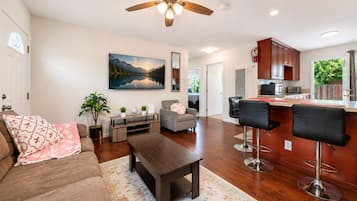 A living room with a sofa, coffee table, TV, and a painting. There is a kitchen with a bar area and a dining table.