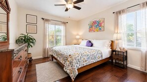 Un dormitorio con cama, ventilador de techo, cómoda, mesita de noche, lámpara y ventana con cortinas.