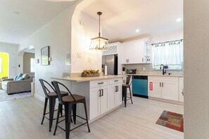 A modern kitchen with white cabinets, a central island, bar stools, and a hanging light fixture. - Hollywood 3BR - Private Pool & Fire Pit (Hollywood)