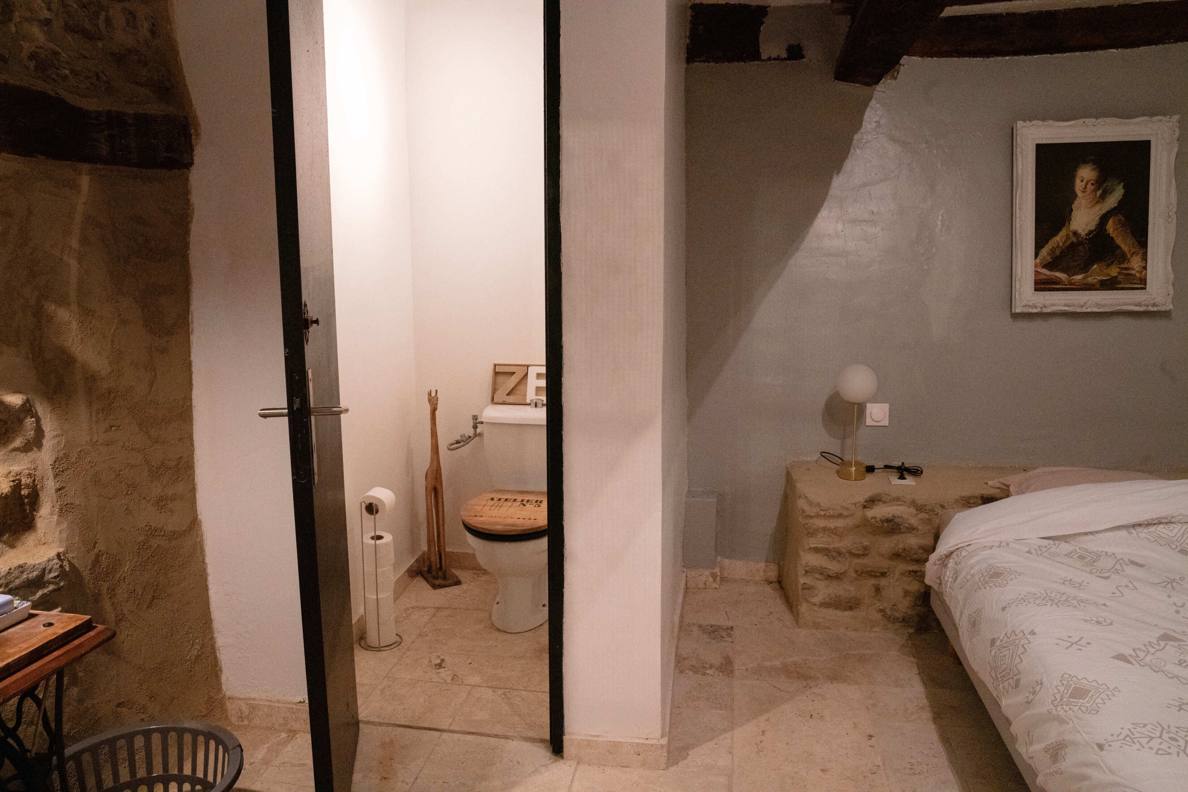 Une pièce avec un mur en pierre, une porte en bois, un lit avec une literie à motifs, une table de chevet avec une lampe et un tableau encadré au mur.
