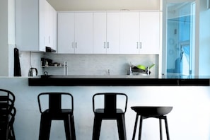 A modern kitchen with white cabinets, a black countertop, and three black bar stools. - Botanika Estate (Jawa Barat)