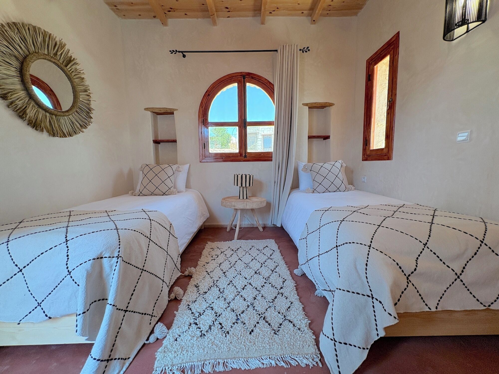 A room with two beds, a round mirror, a window with curtains, and a small table.