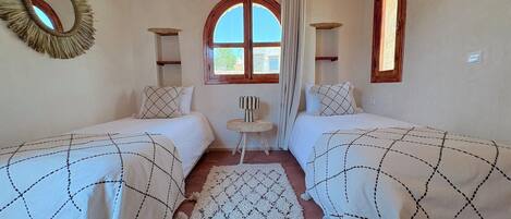 A room with two beds, a round mirror, a window with curtains, and a small table.