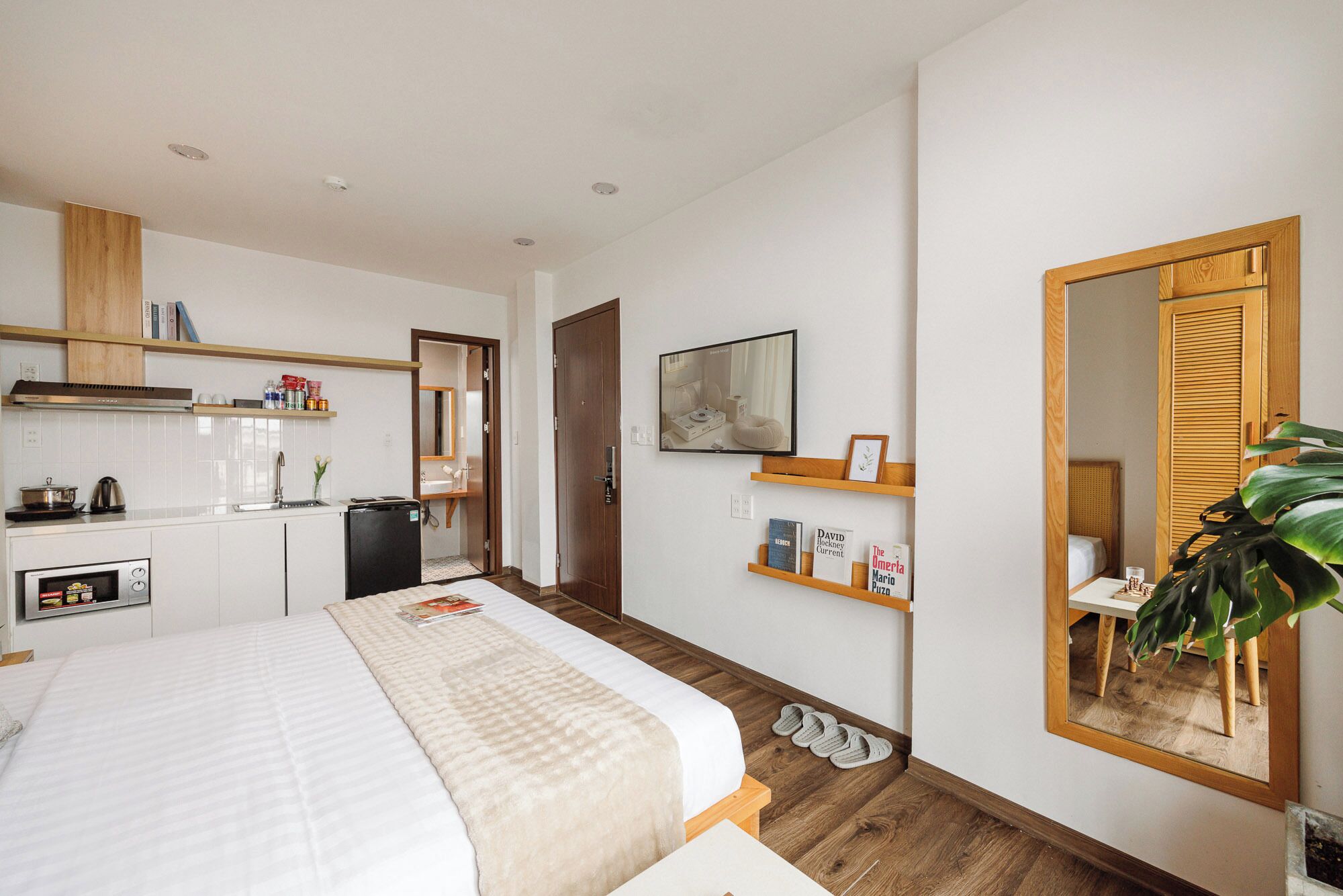 A hotel room with a bed, a kitchenette, a mirror, and a plant.