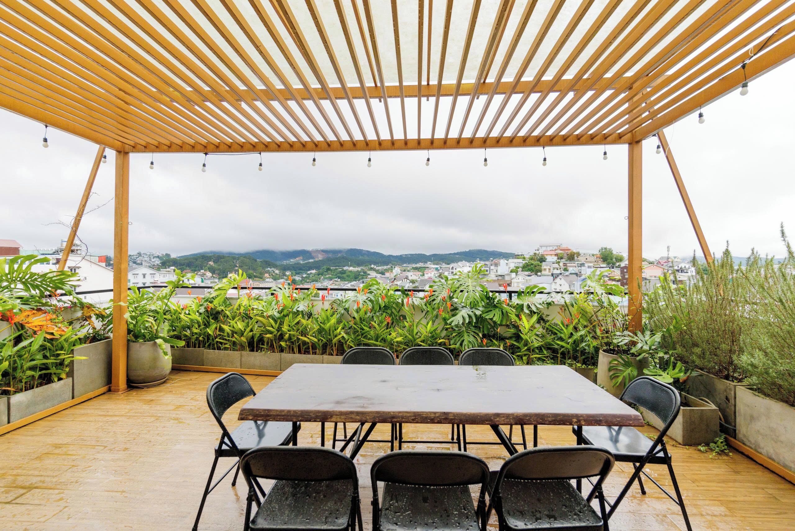 An outdoor patio with a wooden pergola, a table, and chairs, surrounded by potted plants and overlooking a cityscape.