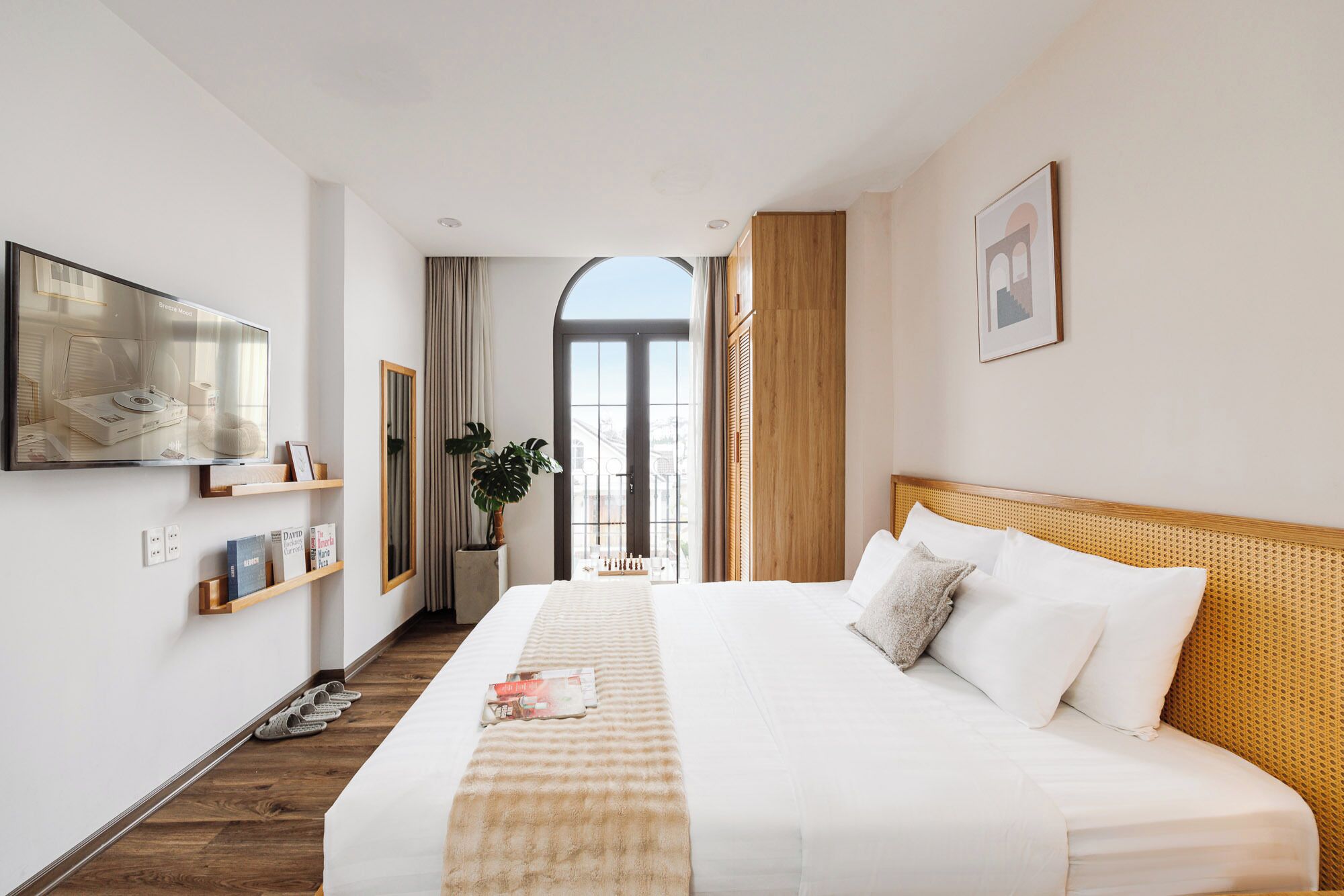 A hotel room with a large bed, a television, a window with curtains, a plant, and a view of the outside.