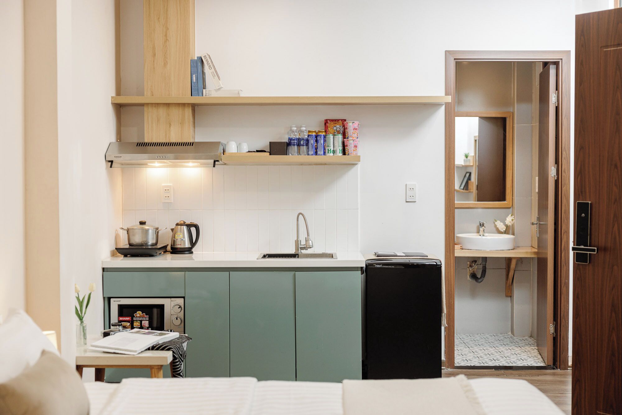 A compact kitchenette with a stove, kettle, and sink, a small table with a laptop and a vase of flowers, and a wooden door leading to a bathroom with a mirror and sink.