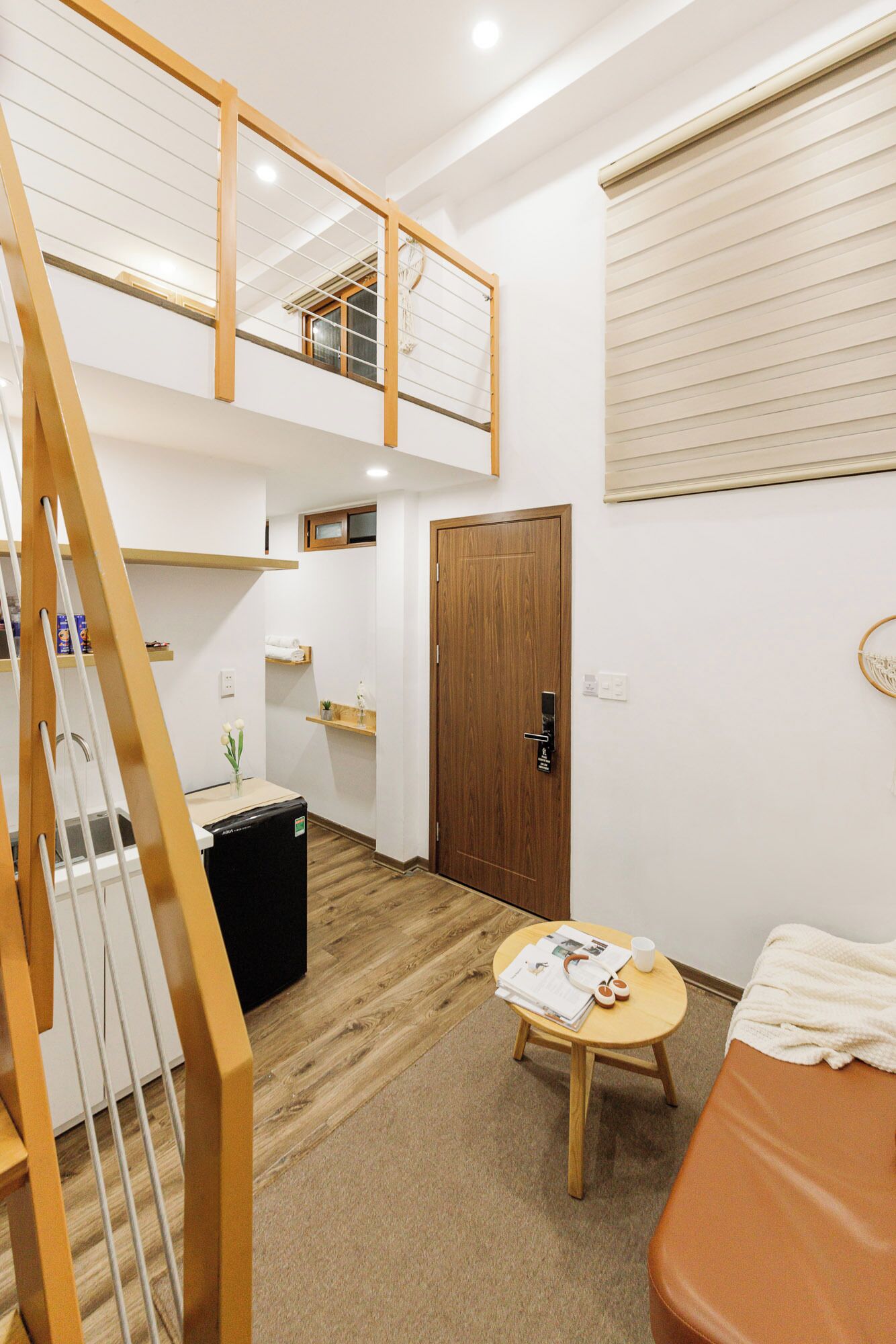 A room with a bunk bed, a small table with a book and a cup, a wooden door, and a window with blinds.
