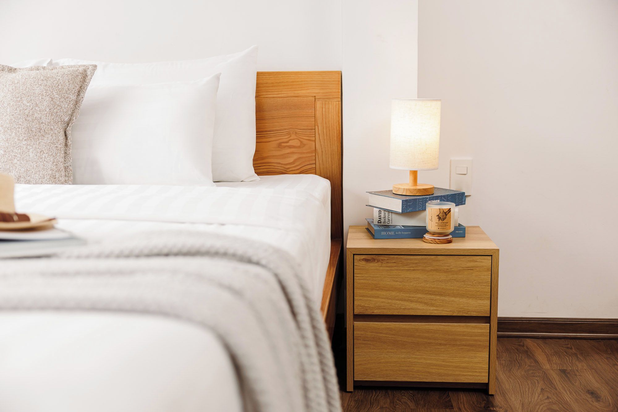 Uma cama bem arrumada com lençóis brancos, cabeceira de madeira e mesa de cabeceira com abajur, livros e uma vela.