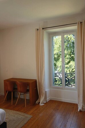A room with a wooden desk and chairs, a large window with curtains, and a view of greenery outside. - 200-square-meter apartment in Villa Tamaris, Aix-les-Bains (Aix-les-Bains)