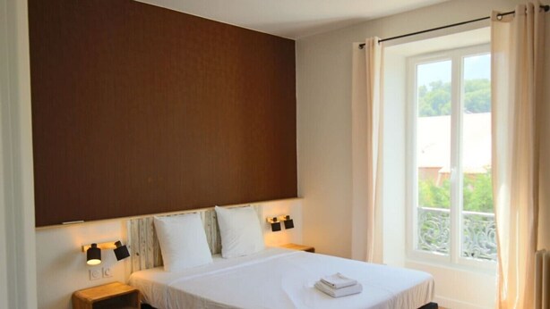 Ein Schlafzimmer mit einem großen Bett, zwei Nachttischlampen, einem Nachttisch, einem Fenster mit Vorhängen und Blick ins Grüne.