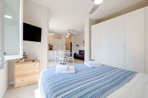 Ein modernes Schlafzimmer mit Bett, einem Holzschrank, einem Fernseher und Blick in die Küche.
